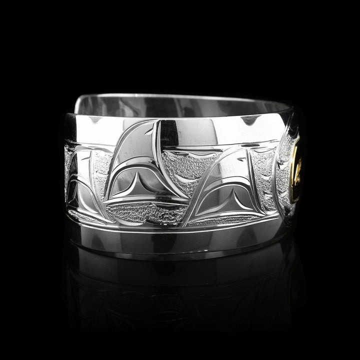 This stunning Orca bracelet is made using sterling silver, 14K gold and has a single, wide band with a gap in the back. There are intricate carvings on the surface of the piece that depicts the Baby Orca, done in gold, in the womb and being watched over by its mother who is depicted in silver and carved on the side of the piece. On the other side of the band, there are depictions dorsal fins in the sea from the Orca pod.