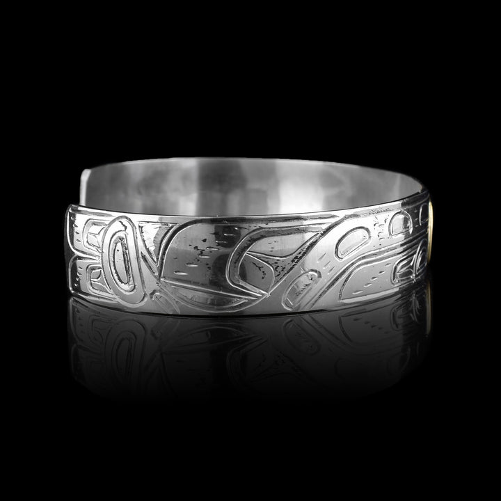This sterling silver and gold bangle bracelet has a single, thin band with carvings on the surface of it depicting the Orca. The head of the legend is carved into a 14K gold oval. On either side of the band, the body of the Orca with a dorsal fin and tail are carved.