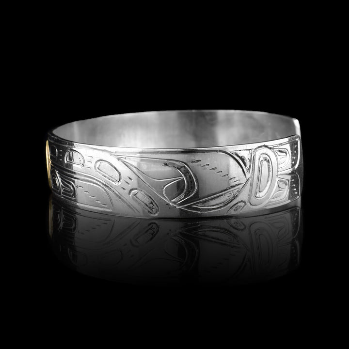 This sterling silver and gold bangle bracelet has a single, thin band with carvings on the surface of it depicting the Orca. The head of the legend is carved into a 14K gold oval. On either side of the band, the body of the Orca with a dorsal fin and tail are carved.