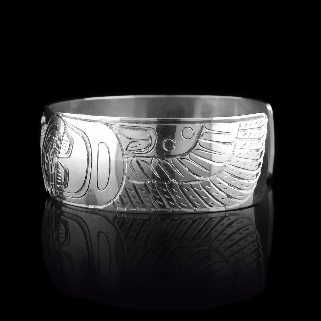 This silver and gold cuff bracelet has a single, wide band with a gap in the back. The band is sterling silver and the Blue Heron is carved into the center of the band. It is depicted in a side view and has a long neck and beak. Its head is done in 14K gold and its wings are depicted on both ends of the piece. The wings have feather details.