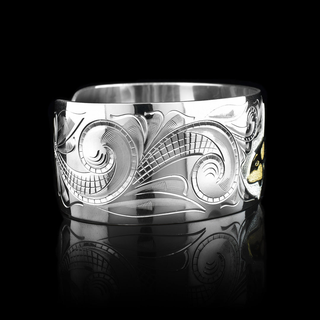 This wide cuff bracelet has a wide, sterling silver band with a gap in the back. There are swirling ocean designs made across the band and a carved orca done in gold.