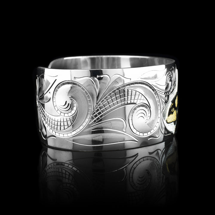 This wide cuff bracelet has a wide, sterling silver band with a gap in the back. There are swirling ocean designs made across the band and a carved orca done in gold.