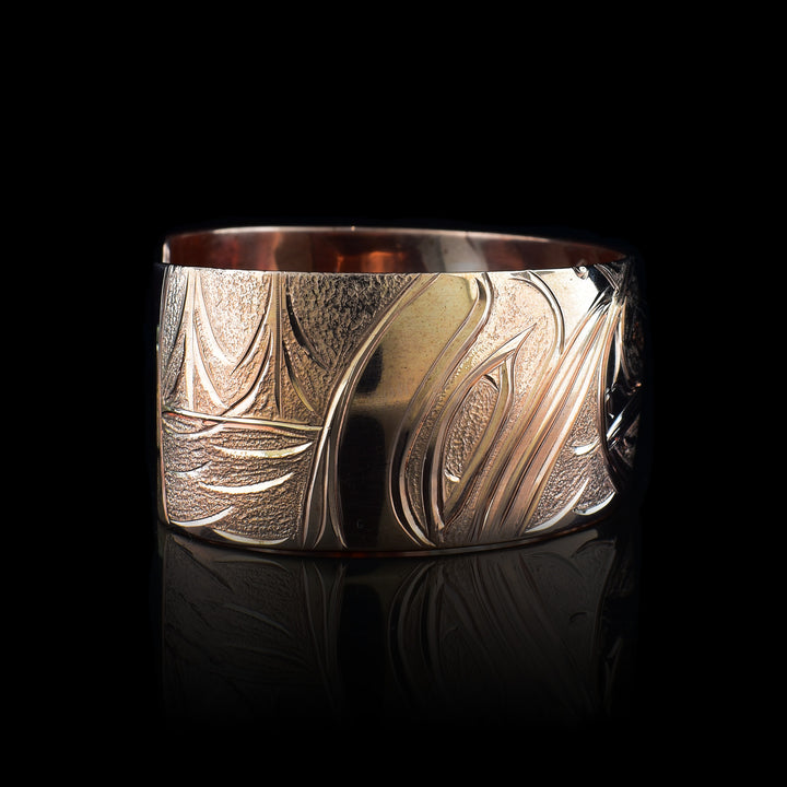 This copper cuff bracelet has a single, wide band with a gap in the back. There is a depiction of the Baby Salmon in its egg carved into the band. In the egg, you can see the eye of the baby Salmon and a fin sticking out on the right side. There are two Salmons depicted on either end of the band watching over the egg and there are forest and river details carved behind them behind them.
