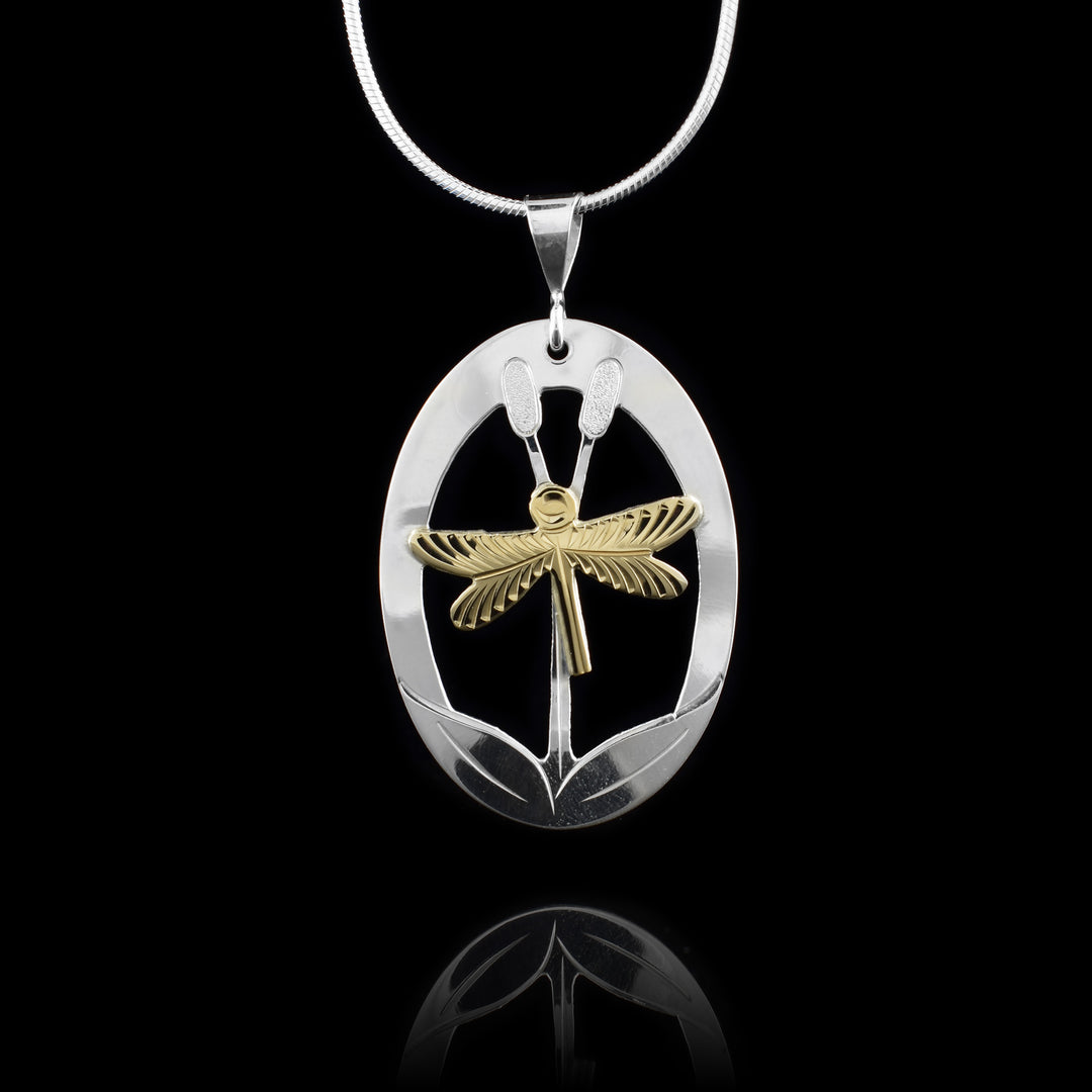 This sterling silver and gold dragonfly pendant is oval shaped and has a cutout designs in the center around a dragon fly. The dragonfly is done in gold and its shown resting on two cattails reeds. There are carvings on the surface detailing the scene.