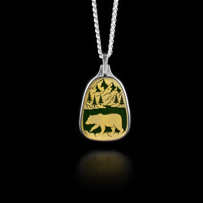 This jade pendant has a 14K gold panel depicting an image of a bear walking along a lake with mountains and trees in the background.