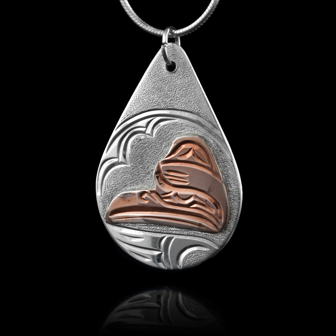 This teardrop pendant necklace is made from sterling silver and copper. There is a depiction of the Raven carved into the piece. The depiction is shown at a side profile and the head of the Raven is done using copper. Feathers, its eye and beak are detailed and there is a wig design carved below the head in the silver.