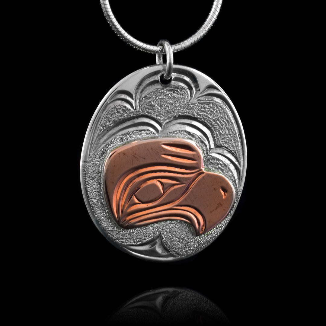This small oval pendant is made from sterling silver and copper. There is a depiction of the Eagle carved into the piece. The depiction is shown at a side profile and the head of the Eagle is done using copper. The eye and the beak are shown with feather details on it and wavy designs in the background on the silver.