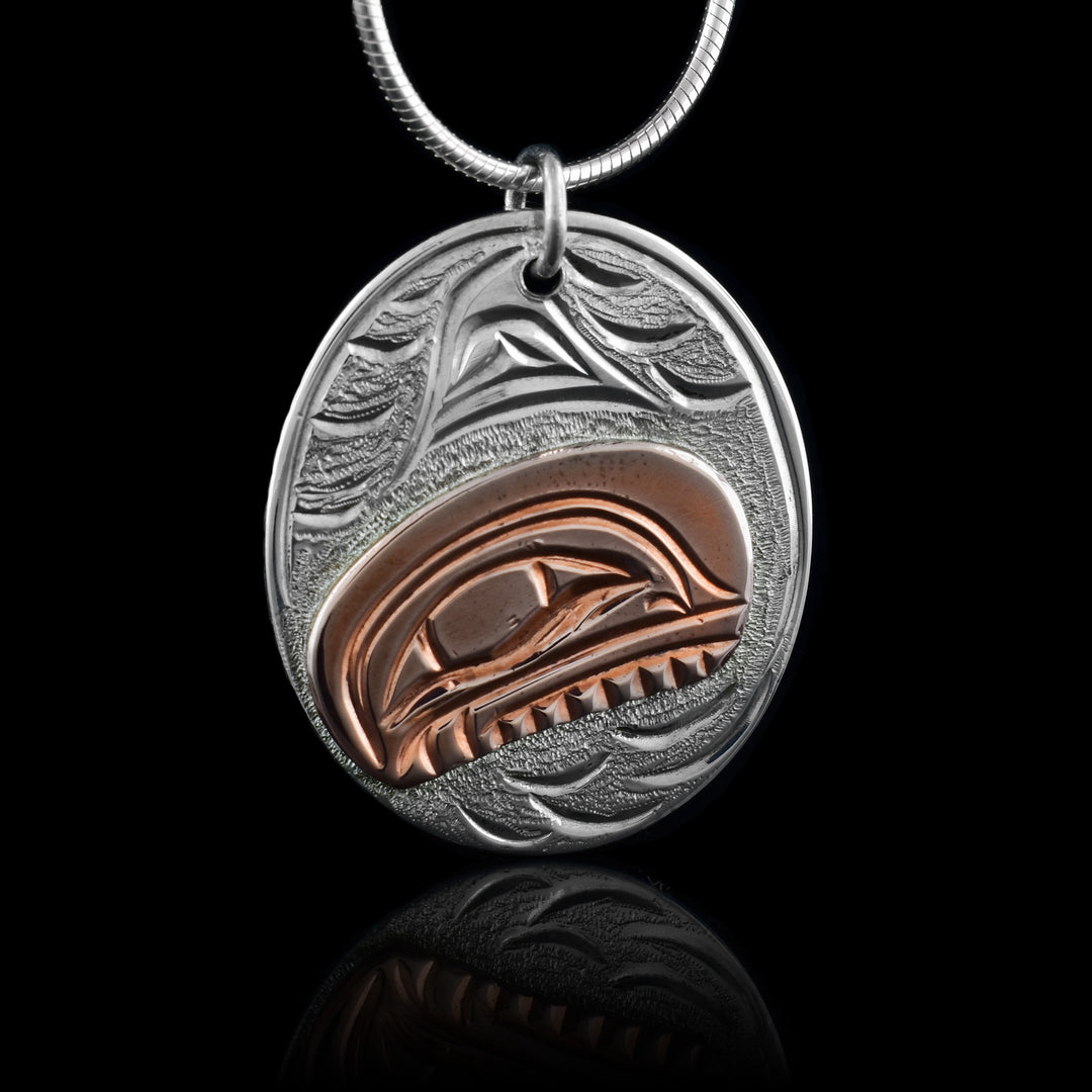 This small oval pendant is made from sterling silver and copper. There is a depiction of the Orca carved into the piece. The depiction is shown at a side profile and the head of the Orca is done using copper. The eye and teeth are shown in the copper with its dorsal fin above it in the silver. There are wave designs in the background of the scene.