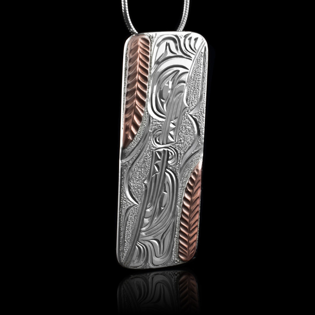 This sterling silver and copper Wolf pendant has a long, rectangle shape with two Wolf heads carved into it. The one at the top is facing down and the other is looking up and their snouts meet in the center. There are copper strips on both ends of the piece that have ridges resembling wheat stalks.