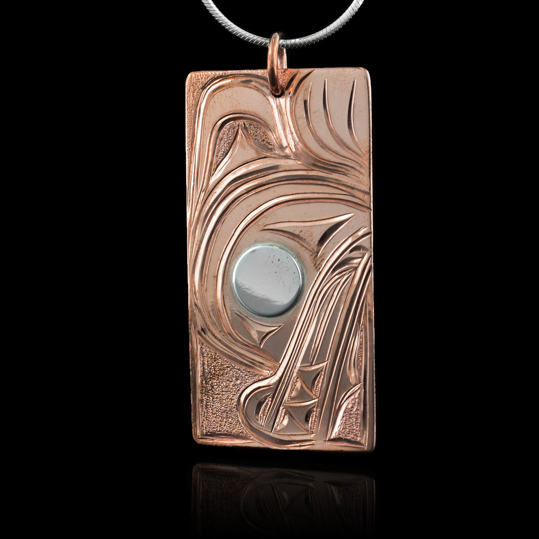This wolf pendant is rectangular in shape and depicts the head of a wolf with a short snout and its teeth showing facing down. Its eye is silver while the rest is copper. Behind the head of the wolf is its large paw.