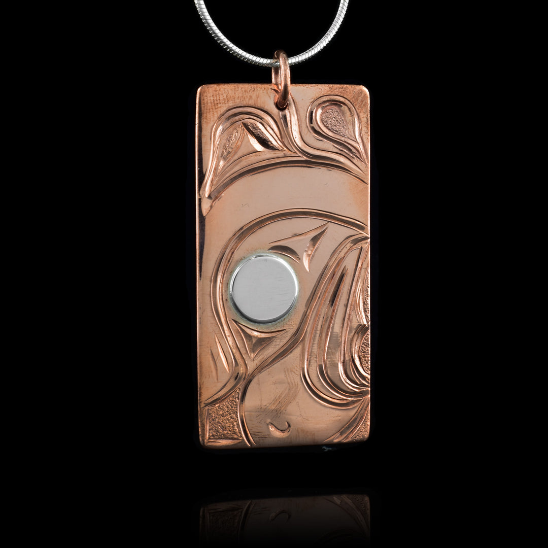 This eagle necklace is rectangular in shape and depicts the head of an eagle facing downward with a feather on its head and a thick, short beak. The eagle's eye is silver while the rest is copper. Behind the head of the eagle is its wing.