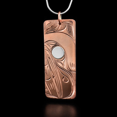 This raven pendant is rectangular in shape and depicts the head of a raven with a large, short beak facing down. Its eye is made from sterling silver while the rest is copper.
