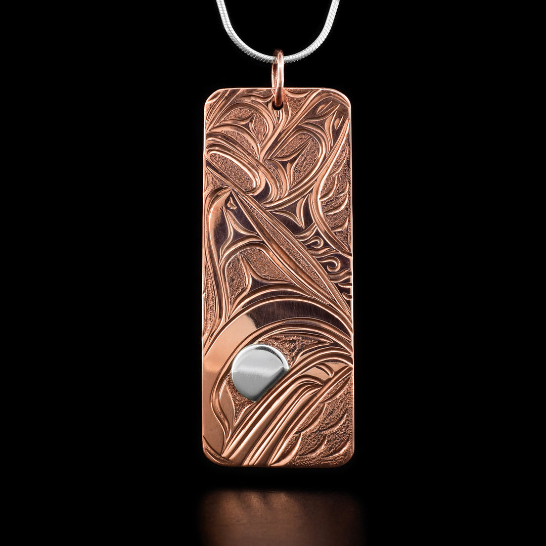 This whale pendant depicts the head of an orca facing downward with its dorsal fin above. The orca's eye is silver while the rest is copper. The orca's large tail has been carved behind its head. Around it are small carvings representing waves.