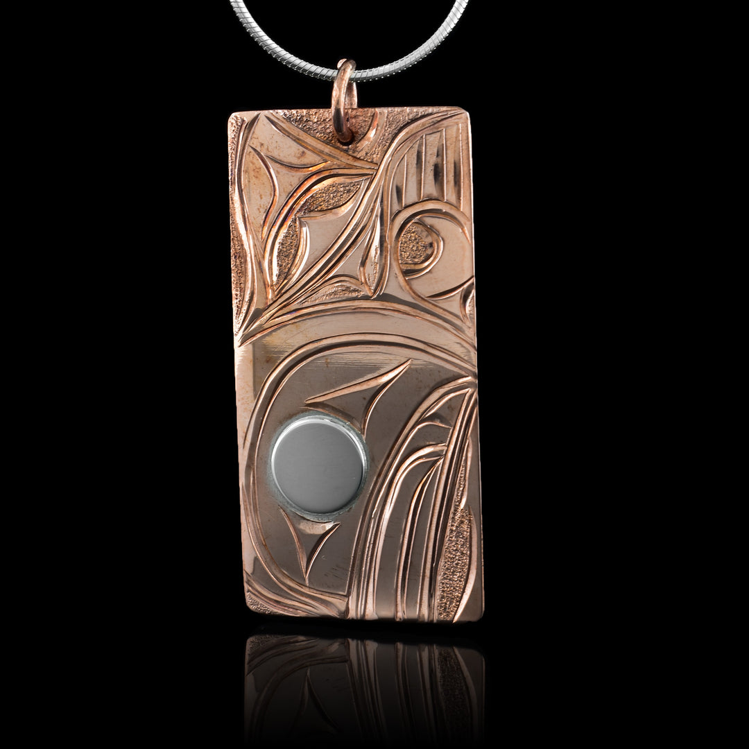 This copper Orca pendant is rectangular in shape and depicts the head of the Orca with a short fin, dorsal fin and its teeth showing facing down. Its eye is silver while the rest is copper. Behind the Orca whale, there are small, textured dimples.