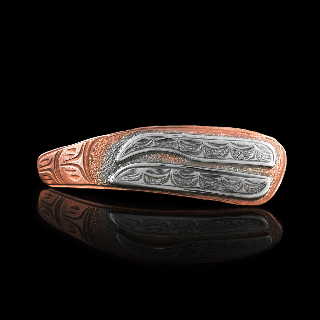 This Orca brooch pin has a long, oblong shape that is made from copper. There are two rows of teeth of the Orca whale that are done in sterling silver and have details carved into them. 
