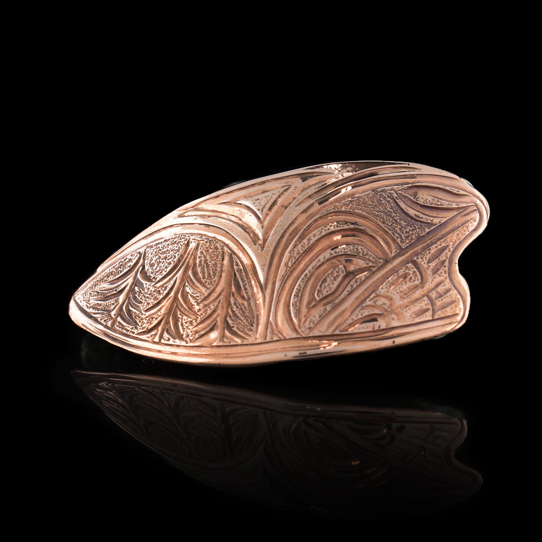 This nature brooch is made from copper and has an irregular shape with rounded side and horizontal length. There is a depiction of trees on one side, repressing a forest, and the head of the Hummingbird is made on the other. The Hummingbird is done at a side profile and has its head tilted upward.