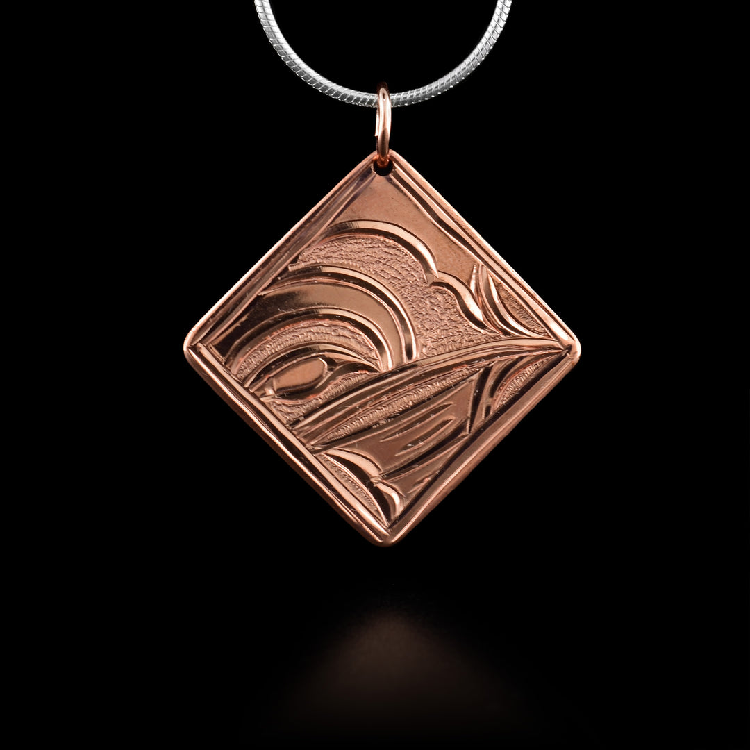 This copper Wolf pendant is square and diamond shaped. There is a thin border around the edges of the piece and the head of the Hummingbird legend is depicted in the center of it. The head is shown at a side profile with its eye, head and beak detailed. There are small dimpled textures in the back ground of the scene.