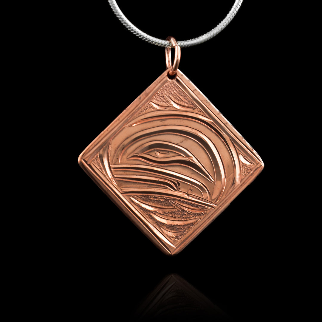 This Orca whale pendant is made from copper and has a square, diamond shape. The head of the Orca is carved into the piece at a side profile. Its eye and mouth are shown and there are wave patterns around the scene.