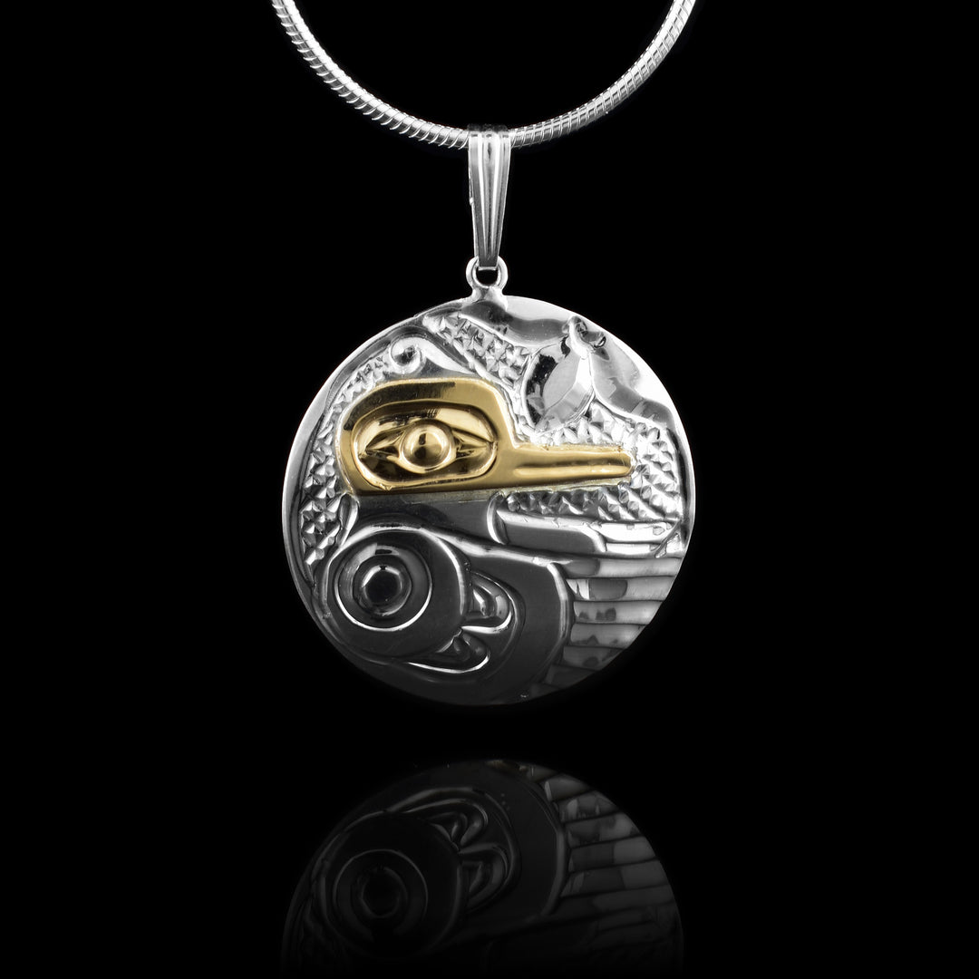 This sterling silver and 14K gold Hummingbird pendant is circle shaped and there is a side profile depiction of the Hummingbird cast into the piece. The head is done in gold with a large eye and thin beak. Its wing is below it and outstretched with designs and feather details included on it. There is a floral pattern at the top of the piece. 
