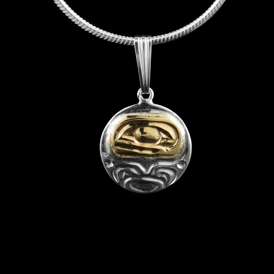 This sterling silver and 14K gold Orca pendant has a very small, circle shape. There is a depiction of the Orca legend cast into the piece. Its large, rectangular head has eye and teeth details with ocean designs around it.