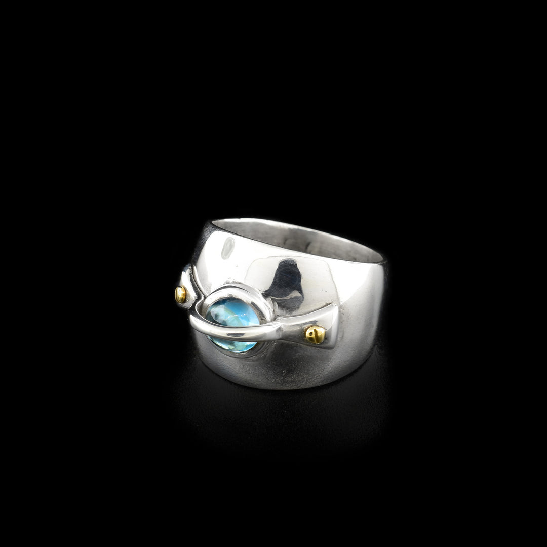 This silver topaz ring is made from sterling silver, 14K gold and blue topaz gemstone. The ring has a wide band with the topaz held in the center and there is a silver band with two 14K gold bold that wraps across the equator of the gem.