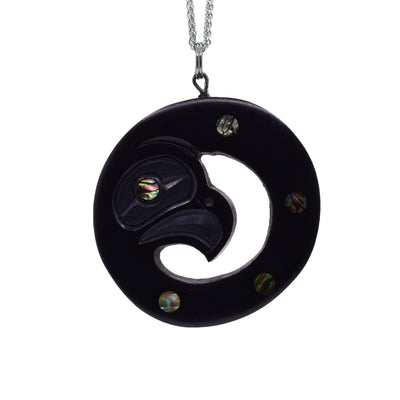 This argillite and abalone pendant is circle shaped and has a cutout in the center around the head of the Eagle. The pendant is made of argillite and has abalone set in the eyes.