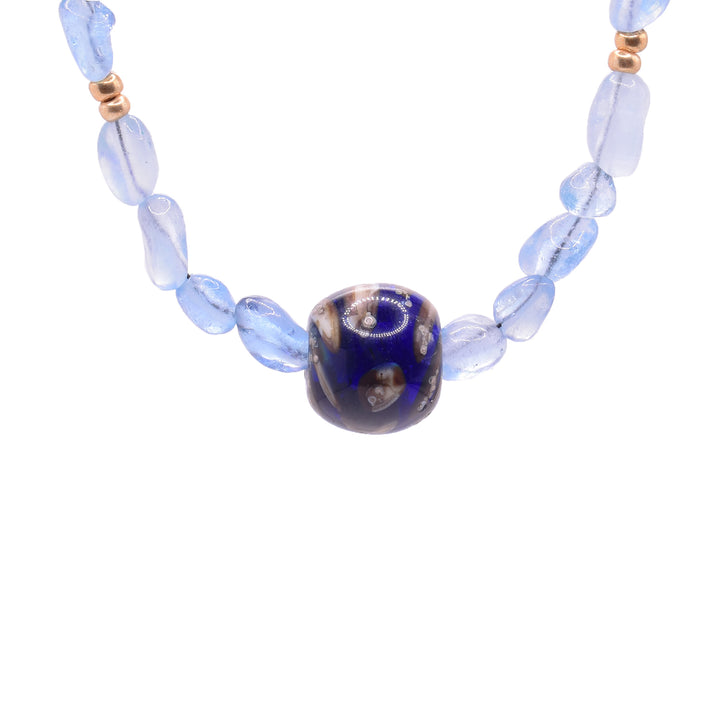 This glass bead necklace has blue glass beads that have rough, oval shapes and run around the piece. There is a large aquamarine bead in the center with light details on it. There are small gold fill beads placed in intervals around the piece.