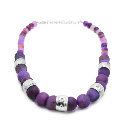 This chunky purple agate necklace has sphere shaped beads that are larger at the bottom of the piece and smaller at the tops of the piece. There are five sterling silver spirti beads featured in the chunky portion of the necklace. There are carvings on the surface of the beads depicting the legends including the Eagle, the Sun, the Moon, the Raven and the Wolf.