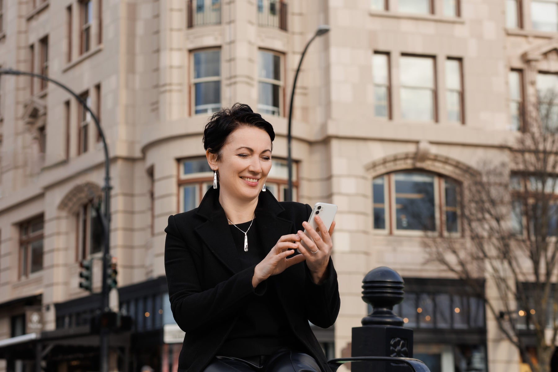 40 to 50-year-old woman in a city wearing sophisticated work attire and Indigenous jewellery looking at an iPhone. Photo is meant to encourage and promote Google Reviews.