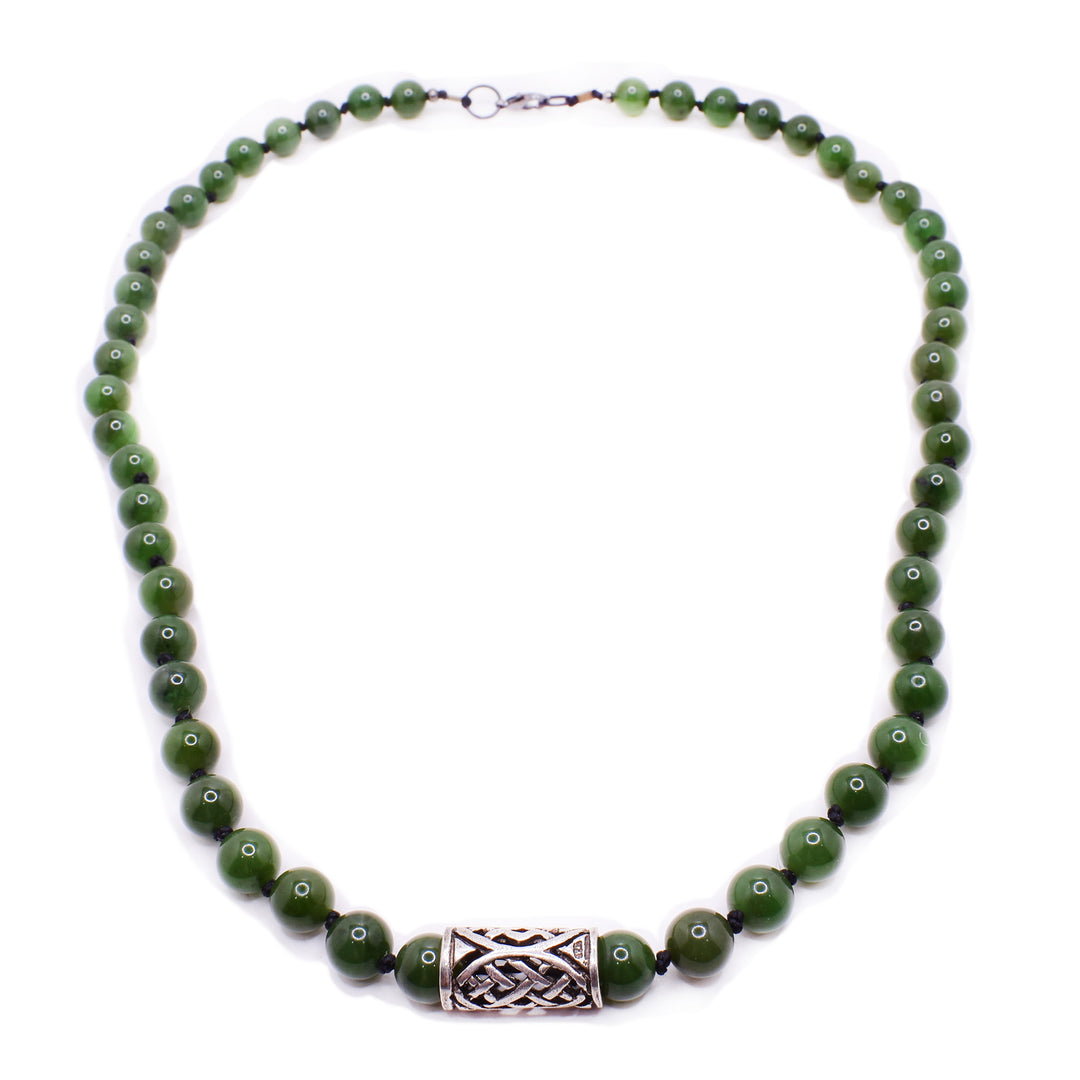 A necklace with round jade beads that has knots in between every bead. There is a sterling silver pendant in the middle. It is tubular in shape, and has a handcarved design on it.