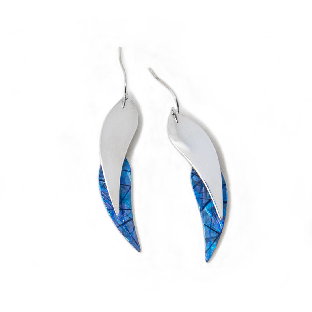 These dangling titanium earrings are made from sterling silver and anodized titanium with leaf shaped hangs. The hangs have a silver leaf with a blue titanium leaf attached to them. There are colorful designs on the surface of the blue metal.
