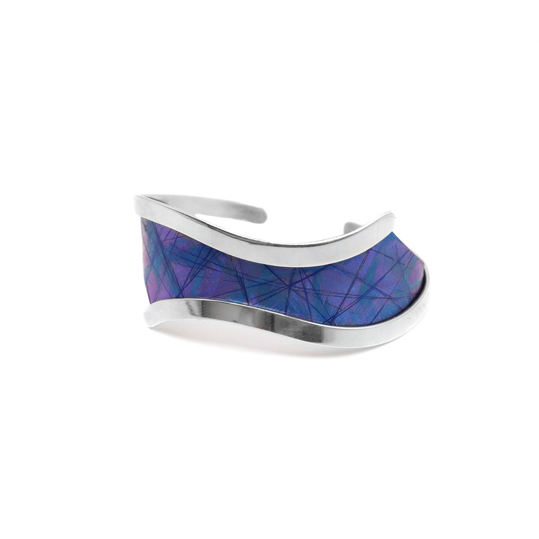 This titanium bracelet cuff has been made with sterling silver and anodized titanium. There is a wide titanium piece at the top of the bracelet and have silver bands on both sides of the piece. The silver bands extend past the wide band and leaves a gap in the back. The titanium is dark blue and purple and has lighter blue designs on the surface of them achieved by anodizing the metal.