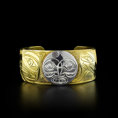This brass bracelet depicts the sun in the center made from silver. The rest of the bracelet is brass and depicts the head of an eagle on the right and the head of a hawk on the left both facing away from the sun.