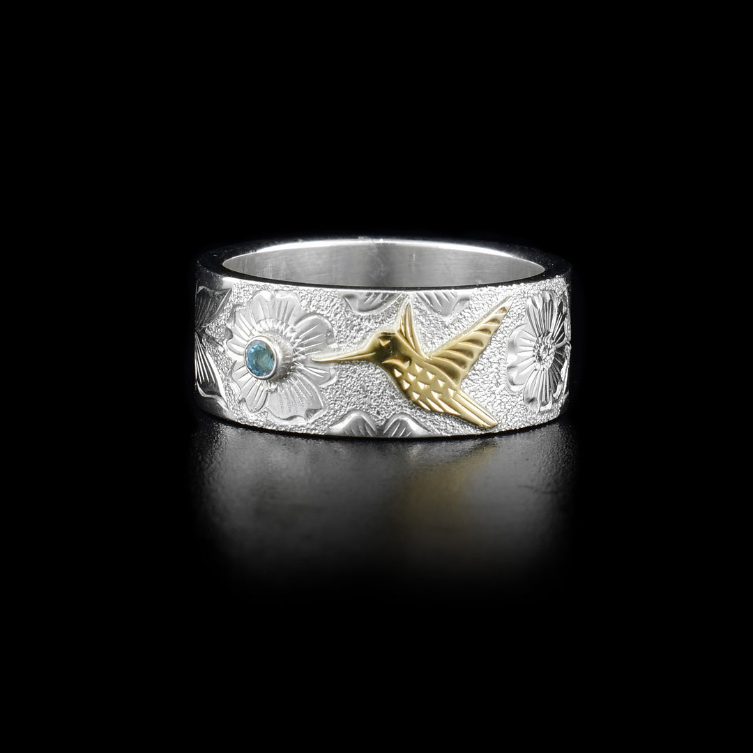 This sterling silver ring is a thick band with a hummingbird and flower carved into it. The humming bird is made from 10K gold and there is a blue topaz gem in the middle of one of the flowers.