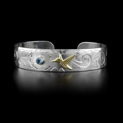 This sterling silver cuff bracelet has carvings depicting flowers and a hummingbird. The hummingbird is made from 10K gold and there is a topaz gemstone in the middle of a flower.