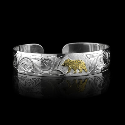 This sterling silver bracelet cuff has a wide band with carvings depicting designs across the band and a bear. The bear is made of 10K gold.