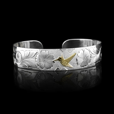 This sterling silver hummingbird bracelet cuff has a wide band with carvings that depict flowers and a hummingbird. The hummingbird is made of 10K gold.