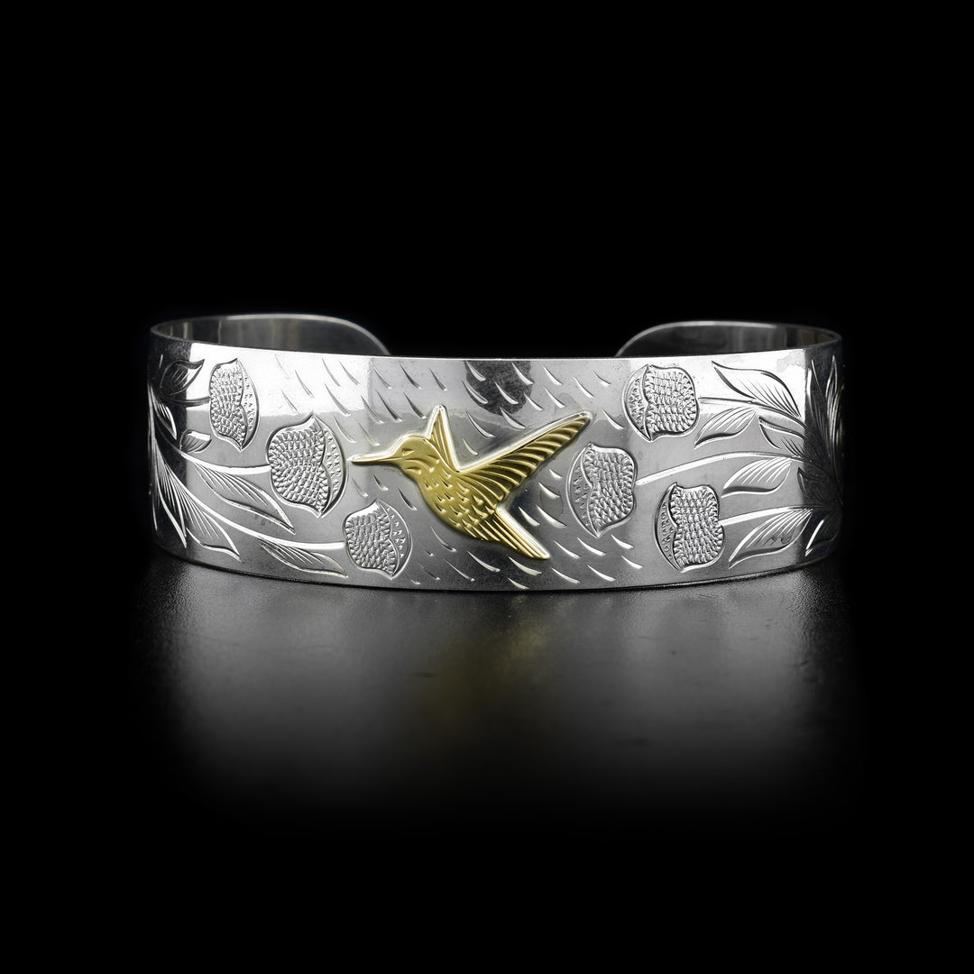 This sterling silver bracelet cuff has a wide band with carvings that depict flowers and a hummingbird. The hummingbird is made of 10K gold.