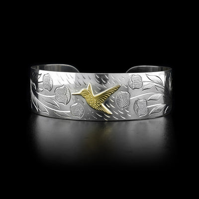 This sterling silver cuff bracelet has a wide band with carvings that depict flowers and a hummingbird. The hummingbird is made of 10K gold.