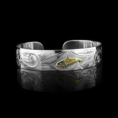 This sterling silver orca bracelet cuff has a wide band with carvings depicting designs across the band and a orca. The orca is made of 10K gold.