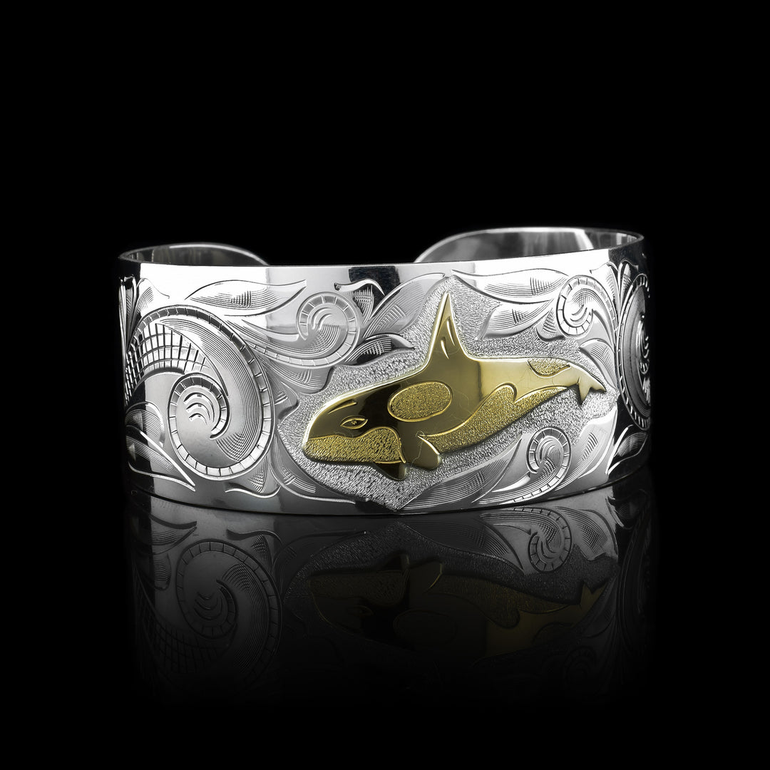 This wide cuff bracelet has a wide, sterling silver band with a gap in the back. There are swirling ocean designs made across the band and a carved orca done in gold.