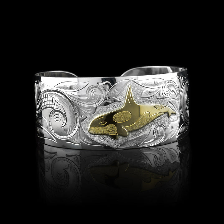 This wide cuff bracelet has a wide, sterling silver band with a gap in the back. There are swirling ocean designs made across the band and a carved orca done in gold.