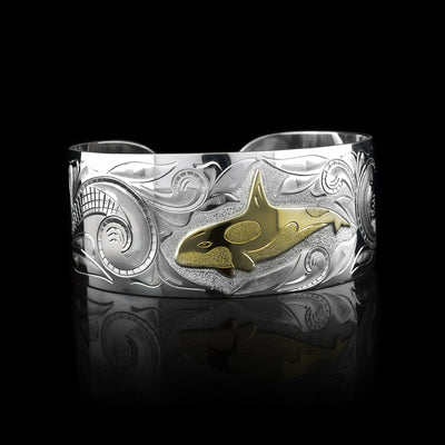 This wide cuff bracelet has a wide, sterling silver band with a gap in the back. There are swirling ocean designs made across the band and a carved orca done in gold.