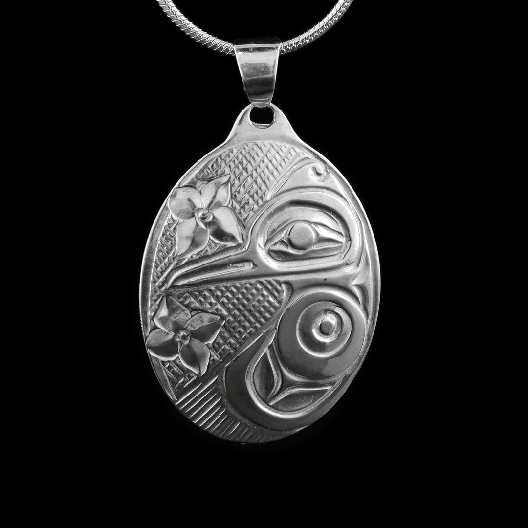 This Hummingbird pendant is made from sterling silver and has a large, oval shape. The Hummingbird is depicted on one side of the piece in a side profile. Its beak extends across the piece and its wing reaches around the bottom of it. There are two flowers depicted across it and there are cross hatching patterns in the background of the portrait.