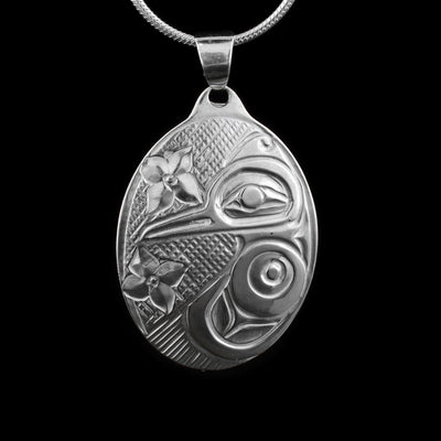 This Hummingbird pendant is made from sterling silver and has a large, oval shape. The Hummingbird is depicted on one side of the piece in a side profile. Its beak extends across the piece and its wing reaches around the bottom of it. There are two flowers depicted across it and there are cross hatching patterns in the background of the portrait.