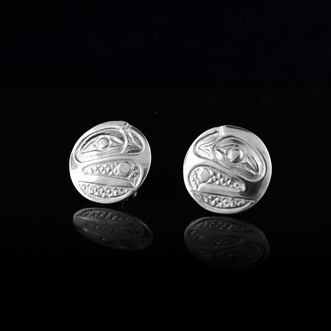 These Circle Stud Earrings are made from sterling silver and have the head of the Raven depicted at a side profile. The eye is large and at the top corner and its beak curves down, is open and is holding a pearl between it. There are cross hatching designs in the background of the portrait.
