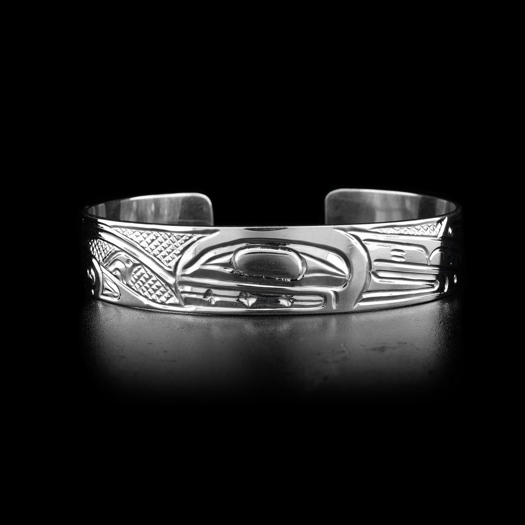 This sterling silver cuff bracelet has a single band with a gap in the back and a depiction of the Orca legend carved into the surface of it. On the right, there are two Orca's depicted in a side profile with large heads and their dorsal fins in view. On the left, there are wave designs with smaller whales breaching from them representing an Orca pod.