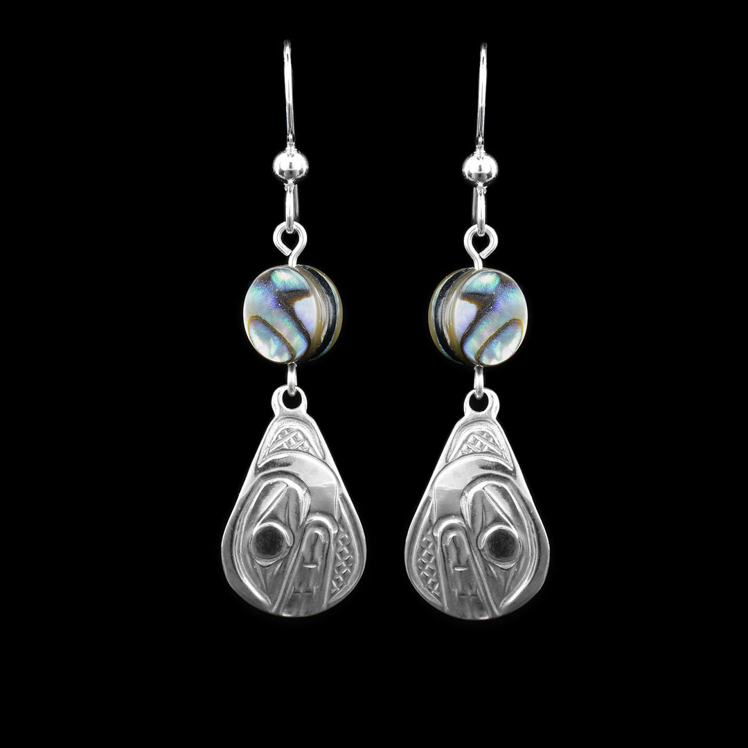 These abalone drop earrings silver have long teardrop shaped hangs made from sterling silver. Below the hooks is a single, round abalone shell charm with the hangs dangling from them. On the hangs, the head of the Orca is shown at a side profile with its teeth and eyes detailed. There are cross hatching designs in the background of the portrait.