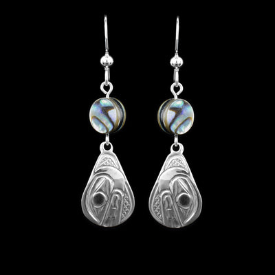 These abalone drop earrings silver have long teardrop shaped hangs made from sterling silver. Below the hooks is a single, round abalone shell charm with the hangs dangling from them. On the hangs, the head of the Orca is shown at a side profile with its teeth and eyes detailed. There are cross hatching designs in the background of the portrait.