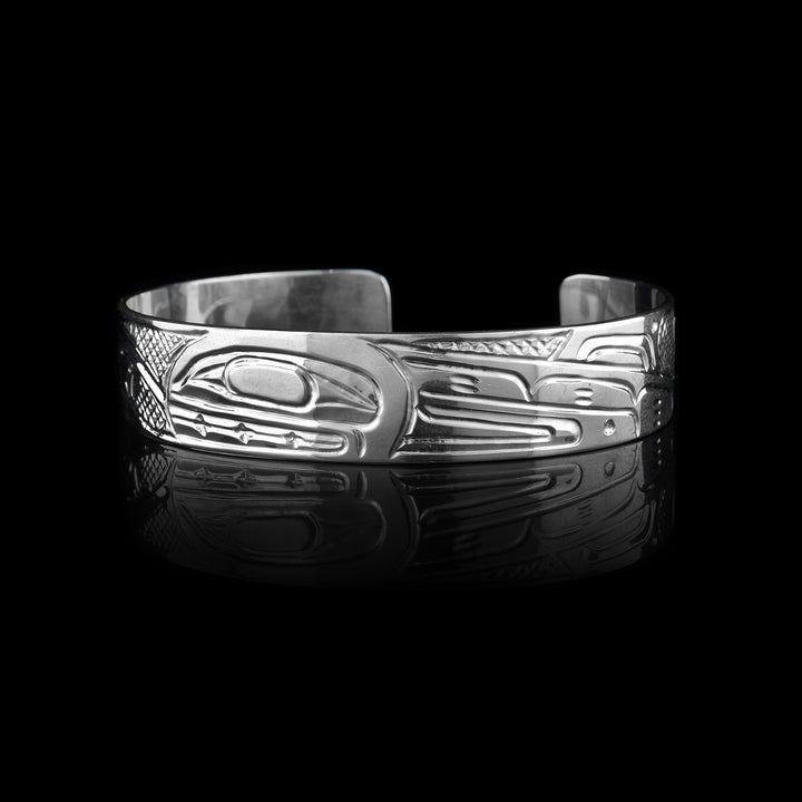 This sterling silver cuff bracelet has a single band with a gap in the back and a depiction of the Orca legend carved into the surface of it. On the right, there are two Orca's depicted in a side profile with large heads and their dorsal fins in view. On the left, there are wave designs with smaller whales breaching from them representing an Orca pod.